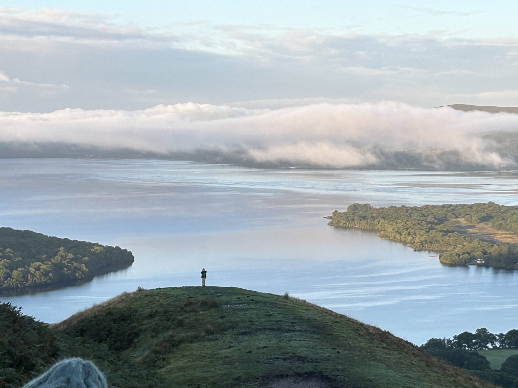 A woman standing alone on a hilltop, looking out over a misty loch